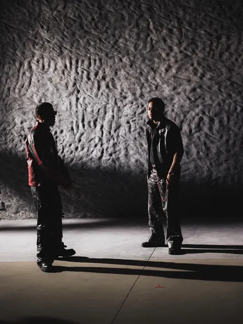 Myke Towers y otra persona de pie frente al muro, cada uno con su sombra