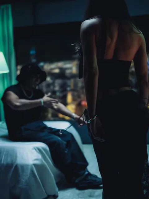 Myke Towers en la cama, hablando con una mujer que está de pie en primer plano